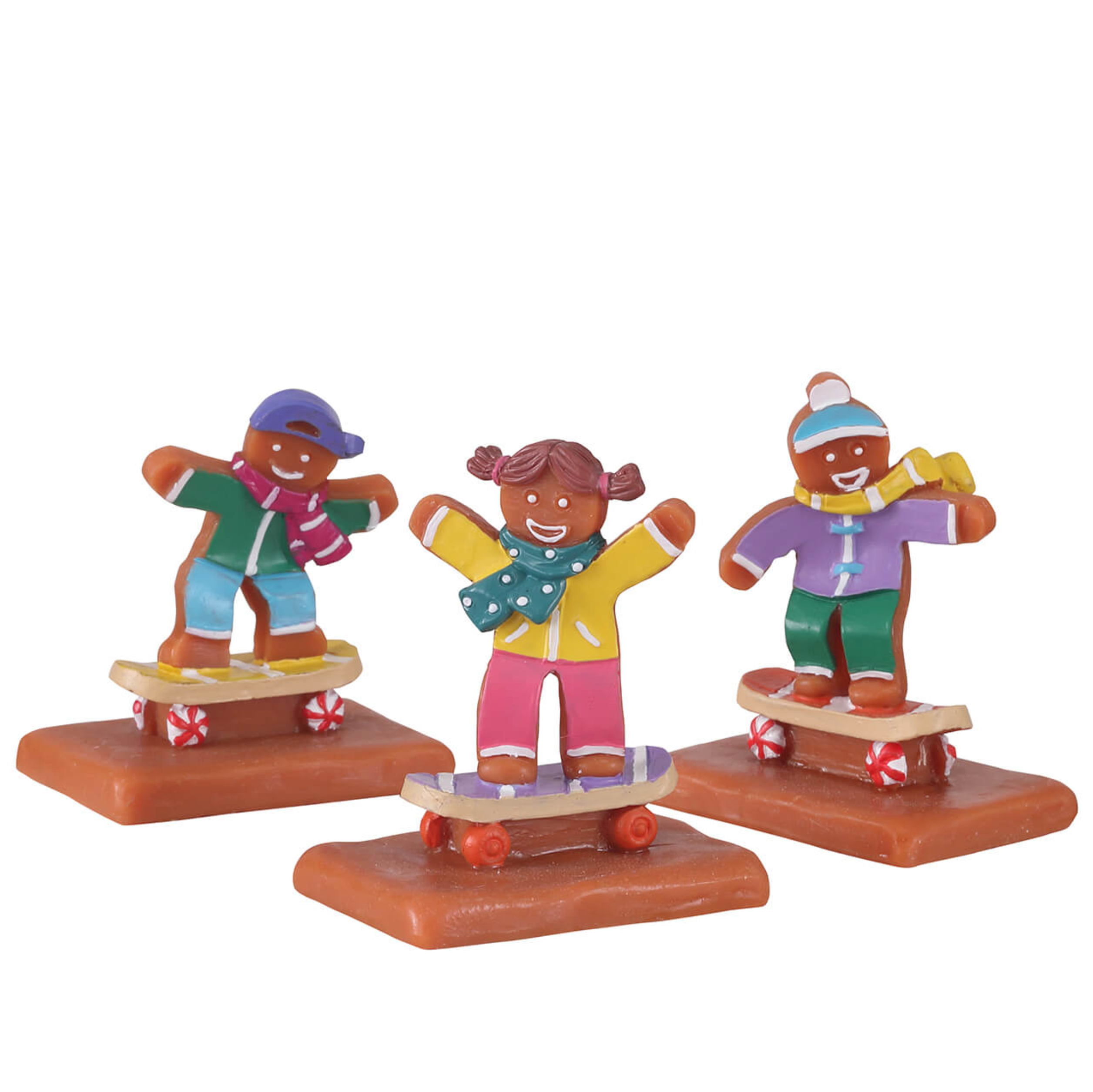 Three Lemax figurines of gindgerbread children on skateboards with colorful outfits on a white background